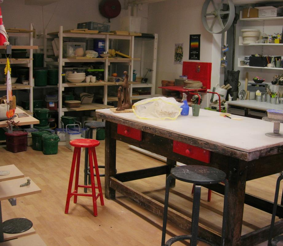 Intérieur de l'atelier : grande table de travail, tabourets et étagères de pièces.